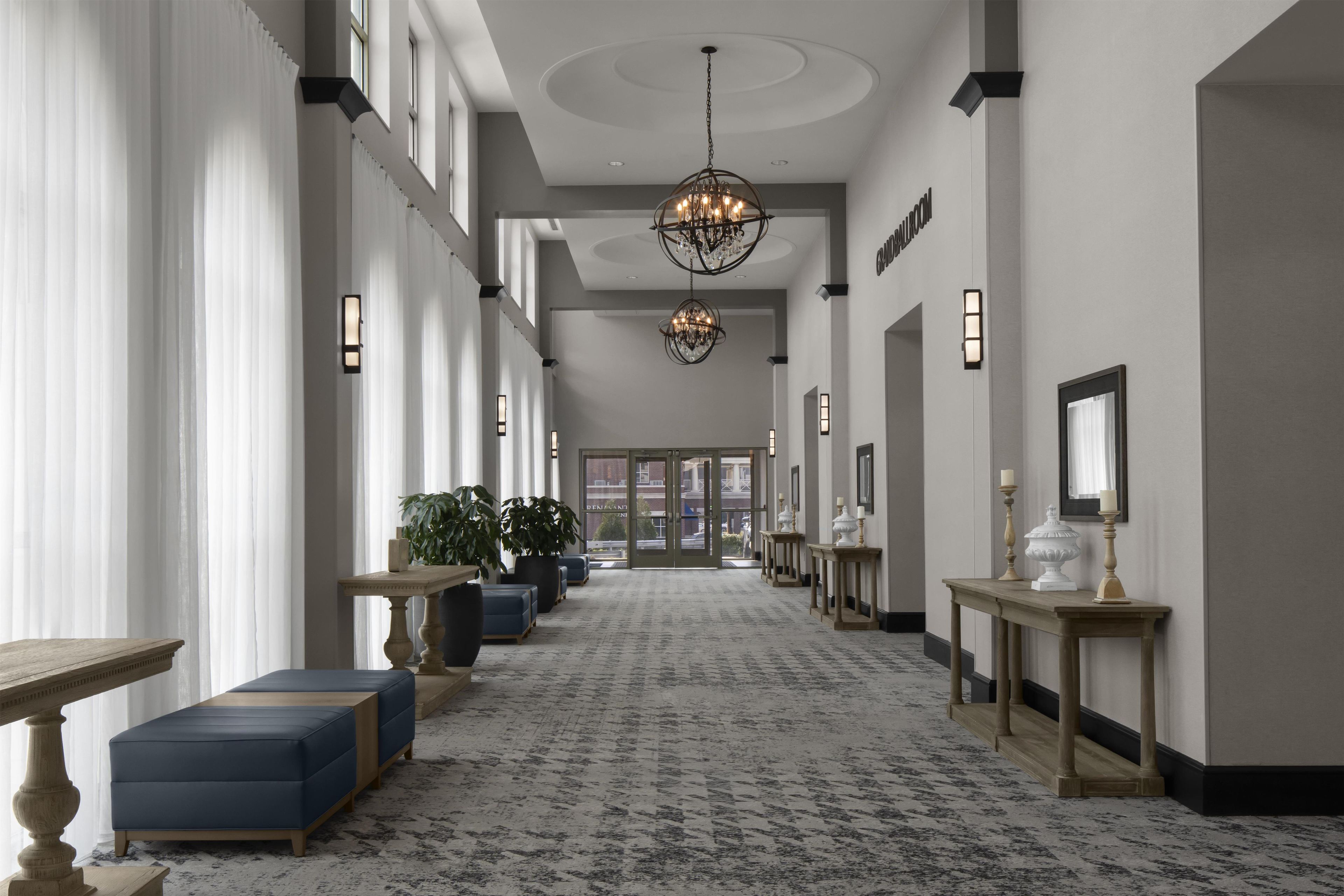 A hotel corridor with a patterned carpet, seating areas, chandeliers, and wall sconces.