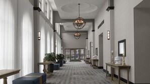 A hotel hallway with a patterned carpet, chandeliers, potted plants, and a reception desk. - Embassy Suites by Hilton Tuscaloosa Alabama Downtown (Tuscaloosa)