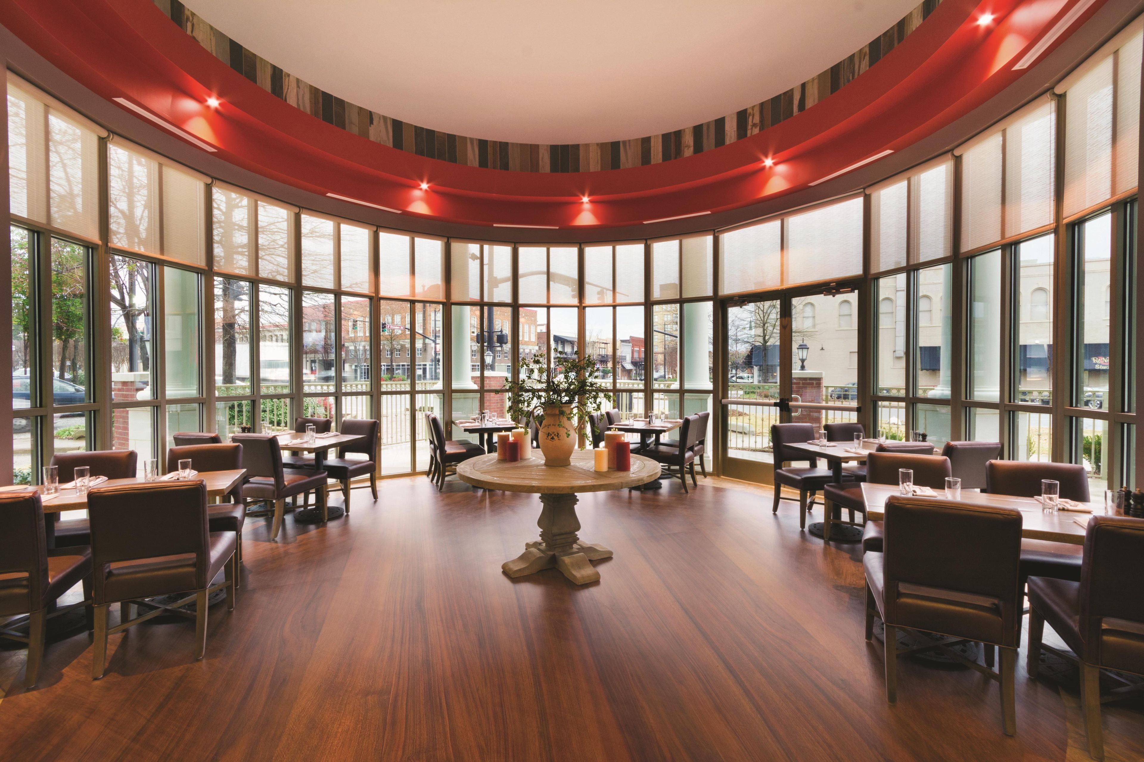 A restaurant with a central round table surrounded by chairs, large windows, and a view of the city.