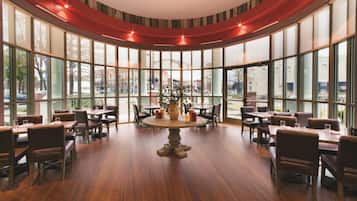 A restaurant with a central round table surrounded by chairs, large windows, and a view of the city.