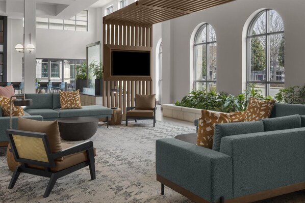 A spacious lobby with a modern design, featuring a large seating area with sofas and chairs, a flat-screen TV mounted on a wooden panel, and large windows offering views of the outside.