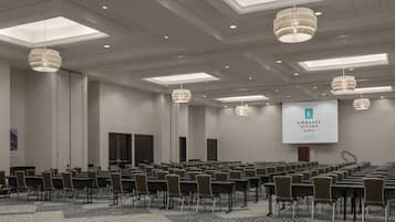 Une salle de conférence avec des rangées de chaises, un grand écran affichant le logo « Embassy Suites by Hilton », et un plafond orné de lucarnes et de lustres.