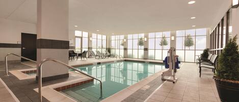 An indoor swimming pool with a tiled floor, a changing room, and a seating area with chairs and a potted plant.