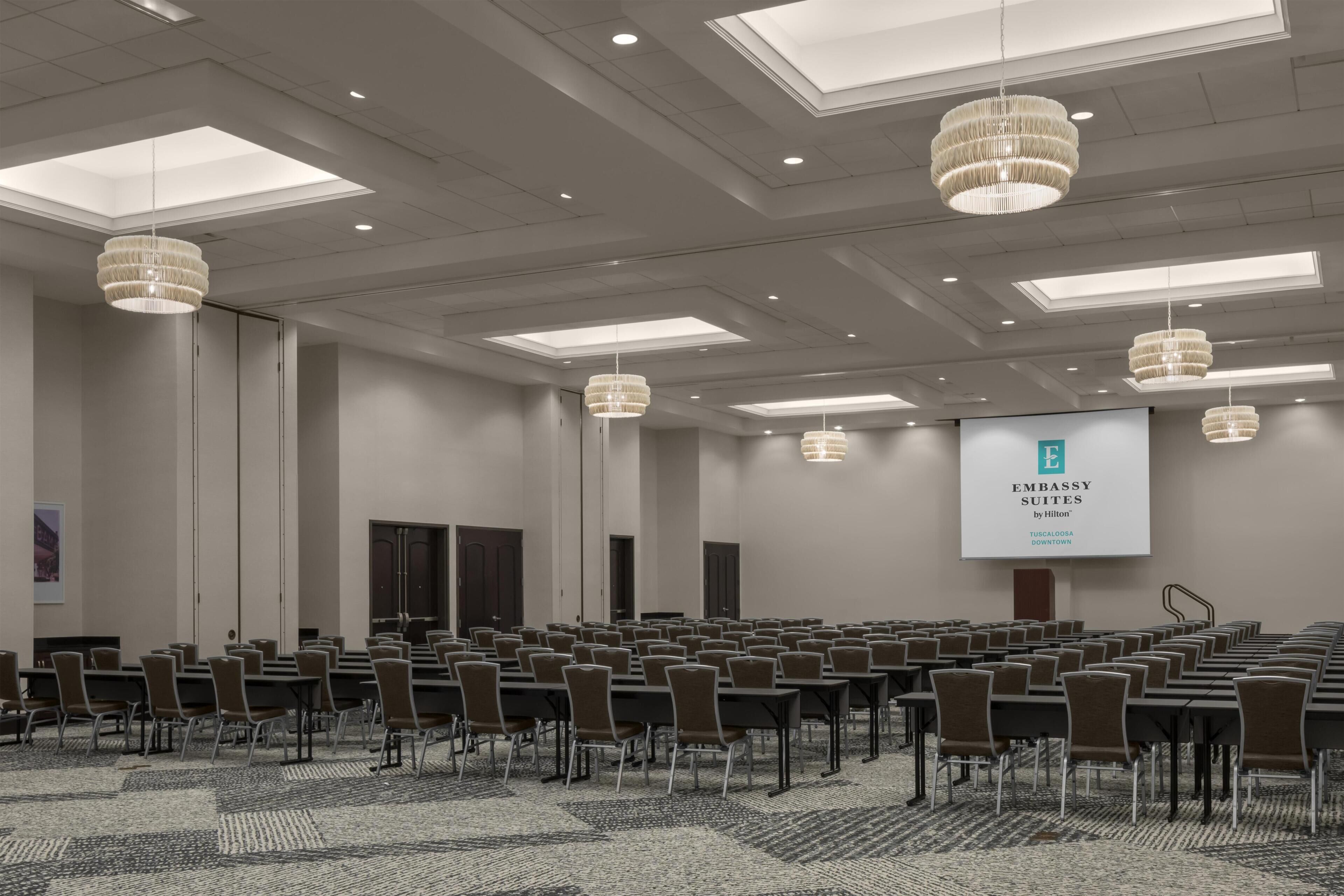 Ein Konferenzraum mit Reihen von Tischen und Stühlen, einem großen Bildschirm, der 'Embassy Suites by Hilton' zeigt, und einer hohen Decke mit Oberlichtern und Kronleuchtern.