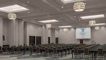 A conference room with rows of tables and chairs, a large screen displaying 'Embassy Suites by Hilton,' and a high ceiling with skylights and chandeliers.