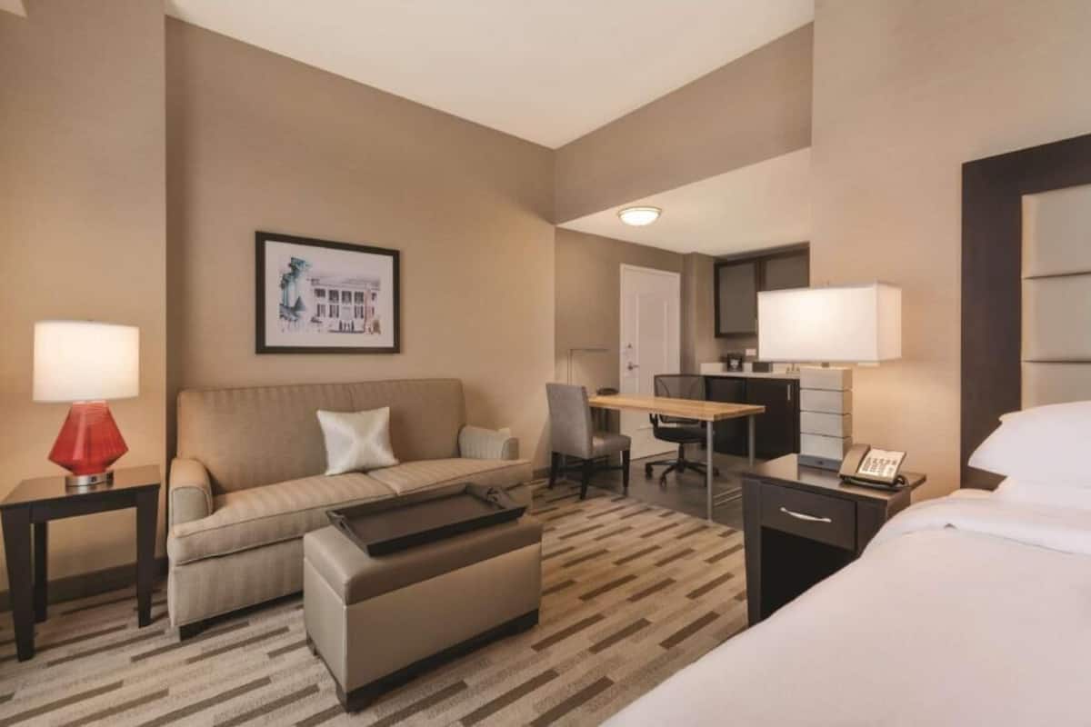 A hotel room with a bed, a sofa, a desk, a chair, a lamp, and a phone.