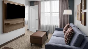 A modern hotel room with a flat-screen TV mounted on the wall, a grey sofa with patterned cushions, a brown ottoman, and a small table.