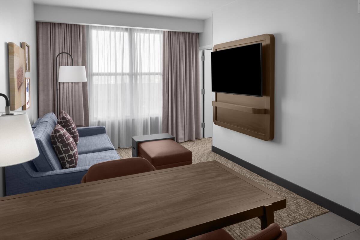 A hotel room with a wooden dining table, a sofa with patterned cushions, a TV mounted on the wall, and a large window with curtains.