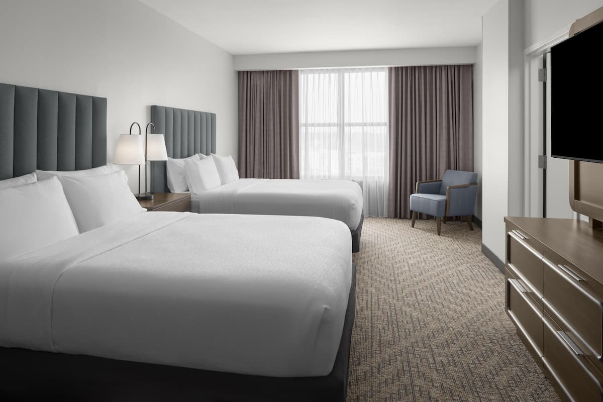 A hotel room with two beds, a desk, a chair, a TV, and a large window with curtains.