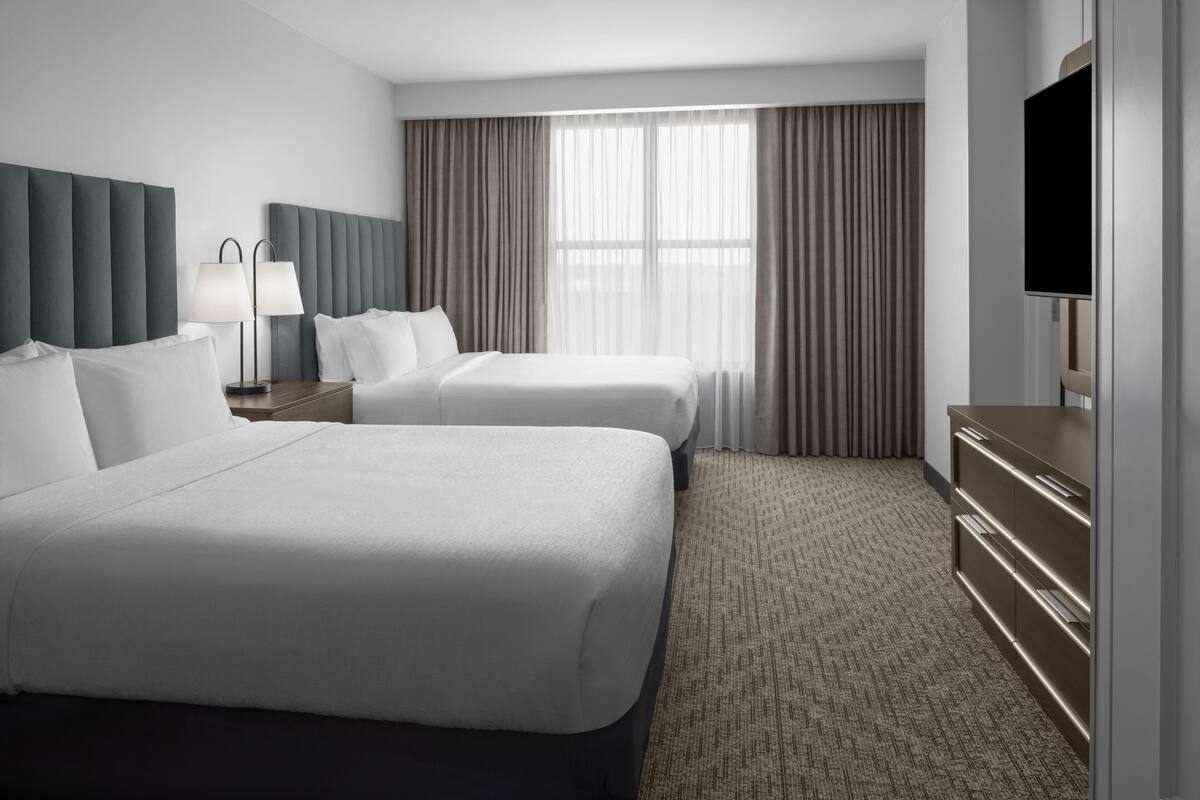A hotel room with two beds, a TV, a desk, and a large window with curtains.