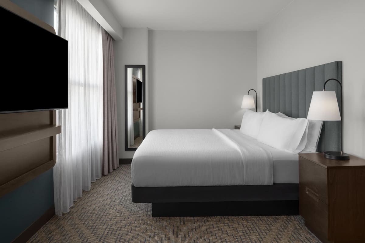 A modern hotel room with a large bed, two bedside lamps, a flat-screen TV, and a wooden nightstand.