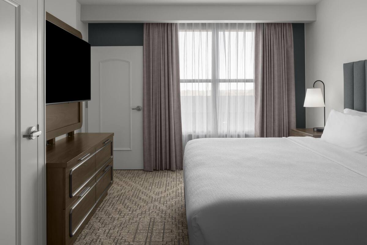 A hotel room with a bed, a TV, a dresser, a lamp, and a window with curtains.