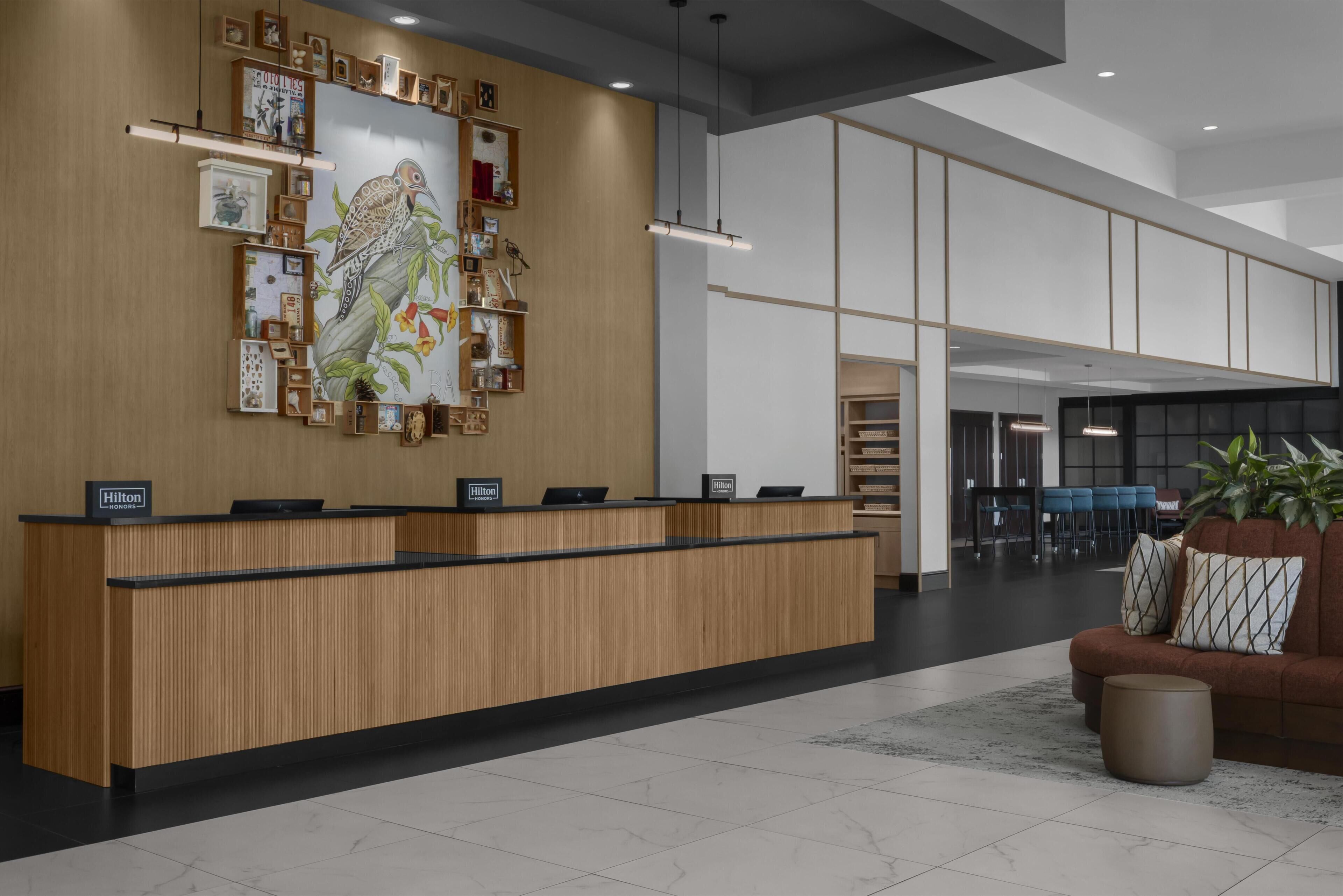 Una recepción de hotel con detalles de madera, un mural grande en la pared y una zona de estar con sofá y planta.