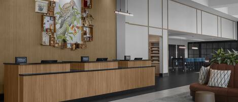 Una recepción de hotel con detalles de madera, un mural grande en la pared y una zona de estar con sofá y planta.