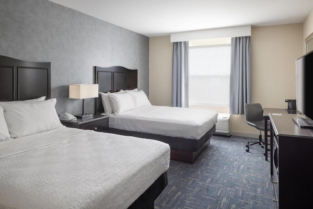 A hotel room with two beds, a desk, a chair, a TV, a lamp, and a window with curtains.