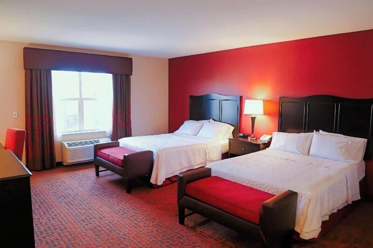 A hotel room with two beds, a desk, a chair, a window with curtains, and a heater.