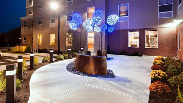 A modern building with a courtyard featuring a central sculpture, a brick facade, and a well-lit entrance. - Hampton Inn Lewiston-Auburn (Lewiston)