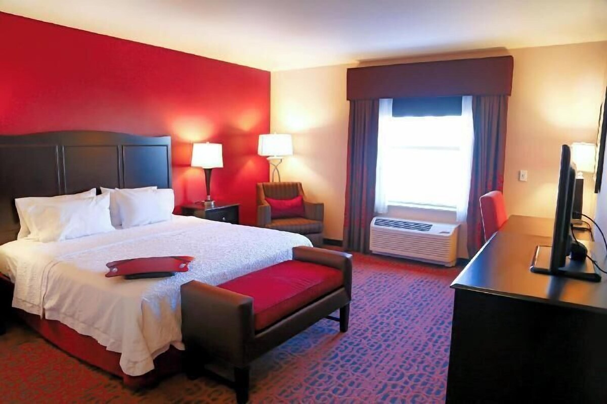 A hotel room with a bed, a desk, a chair, a lamp, and an air conditioning unit.