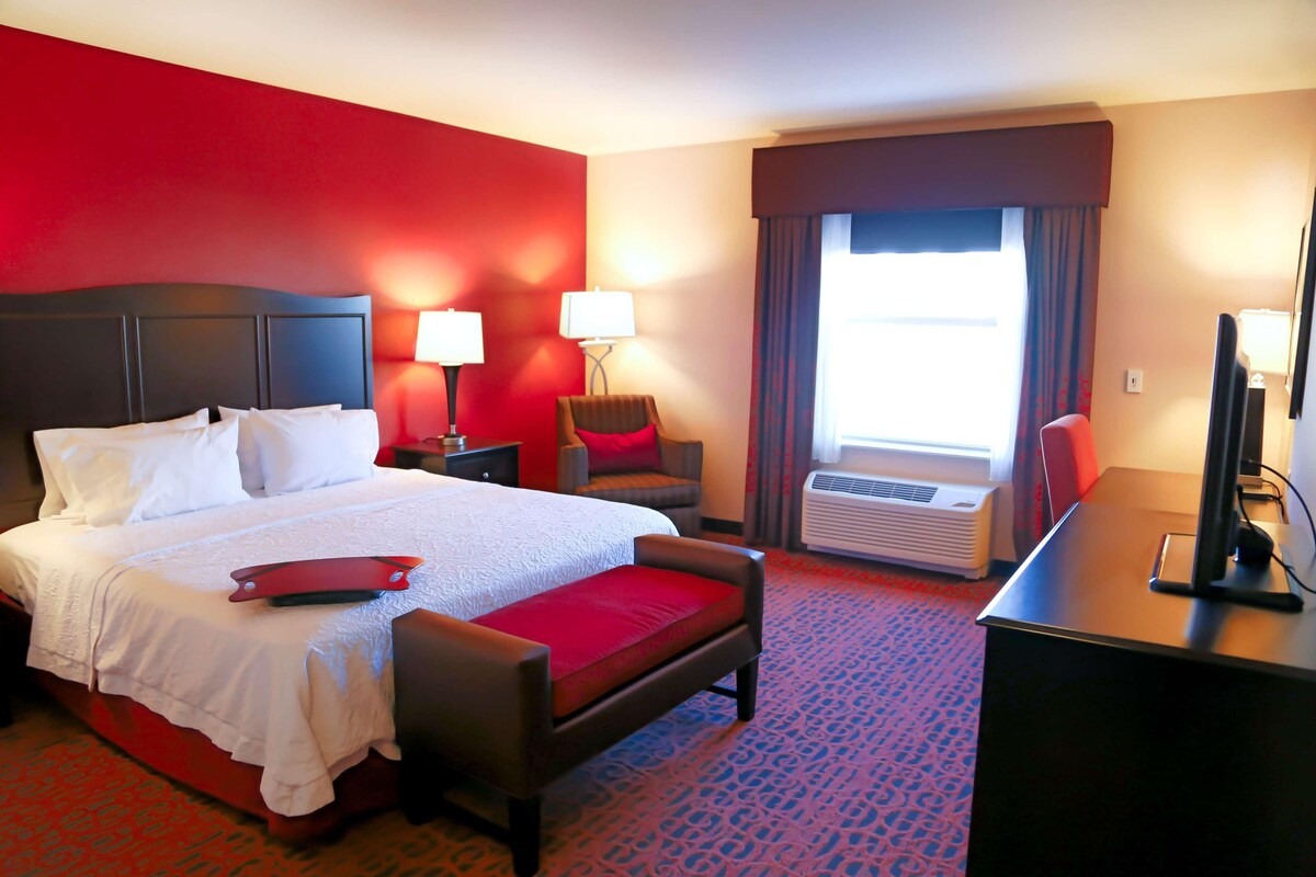A hotel room with a bed, a desk, a chair, a lamp, and an air conditioning unit.