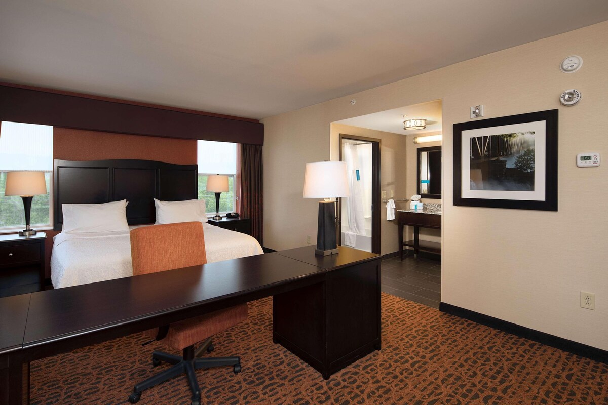 A hotel room with a bed, desk, and chair, a bathroom with a mirror, and a framed picture on the wall.