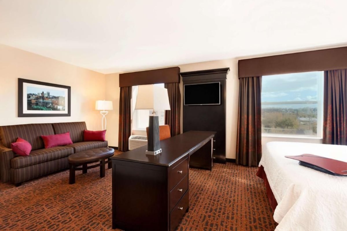 Ein Hotelzimmer mit Bett, Schreibtisch, Sofa, Lampe, Fernseher und Fenster mit Blick.