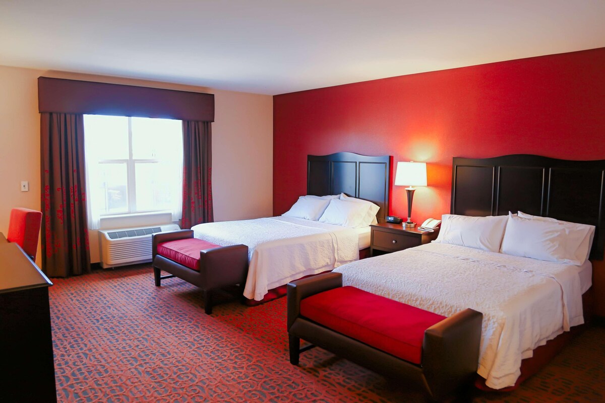 A hotel room with two beds, a desk, a chair, a lamp, and a window with curtains.