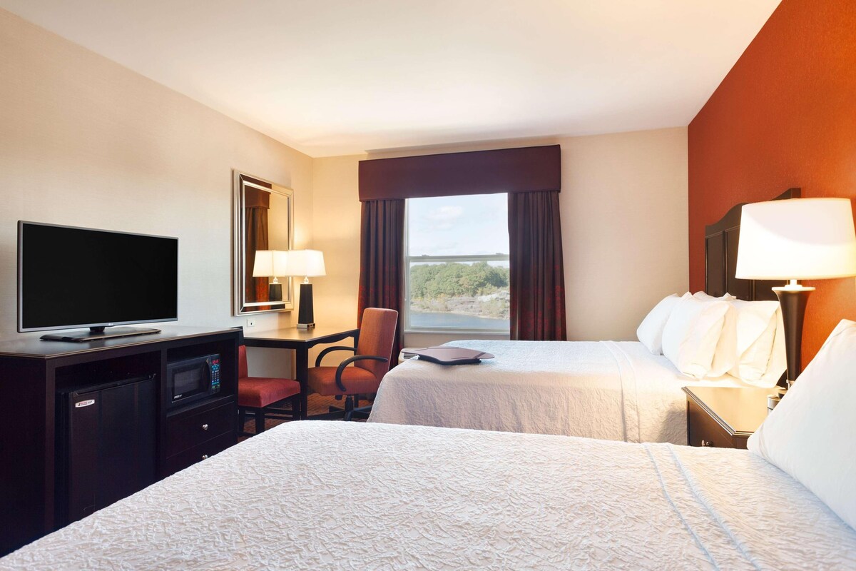 A hotel room with two beds, a desk with a TV, a chair, a desk lamp, and a window with a view.