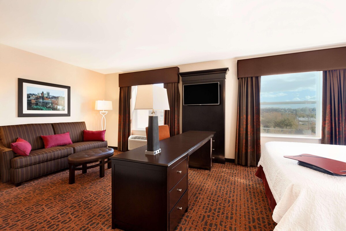 A hotel room with a sofa, a desk, a bed, a TV, and a window with a view.