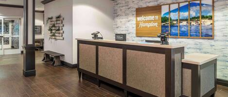 A reception desk with a 'Welcome to Hampton' sign, a wooden bench, and a wall with a scenic view.
