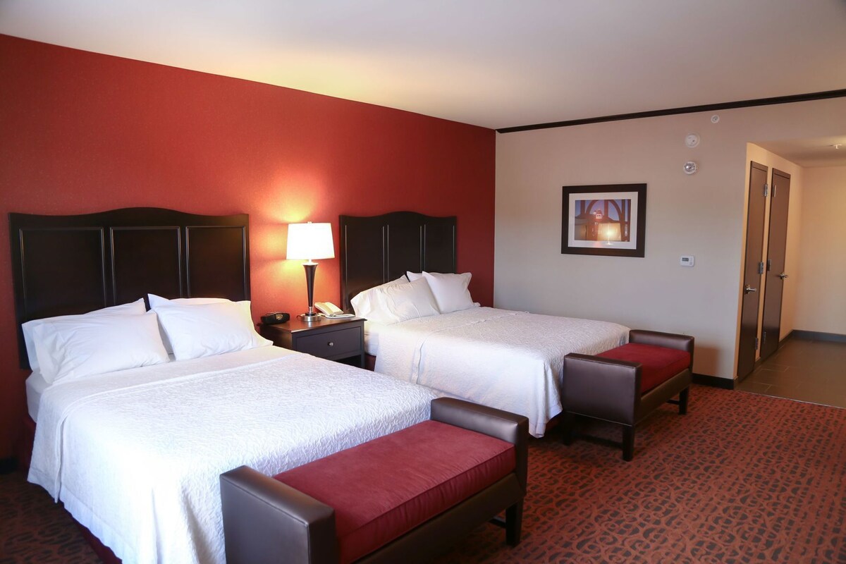 A hotel room with two beds, a nightstand with a lamp, a framed picture on the wall, and a door.