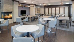 A spacious dining area with multiple round tables and chairs, a stone fireplace, and a flat-screen TV mounted on the wall. - Hampton Inn Lewiston-Auburn (Lewiston)