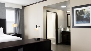 A hotel room with a bed, bedside table, lamp, and a desk. There is a bathroom visible with a mirror, sink, and towel rack. - Hampton Inn Lewiston-Auburn (Lewiston)