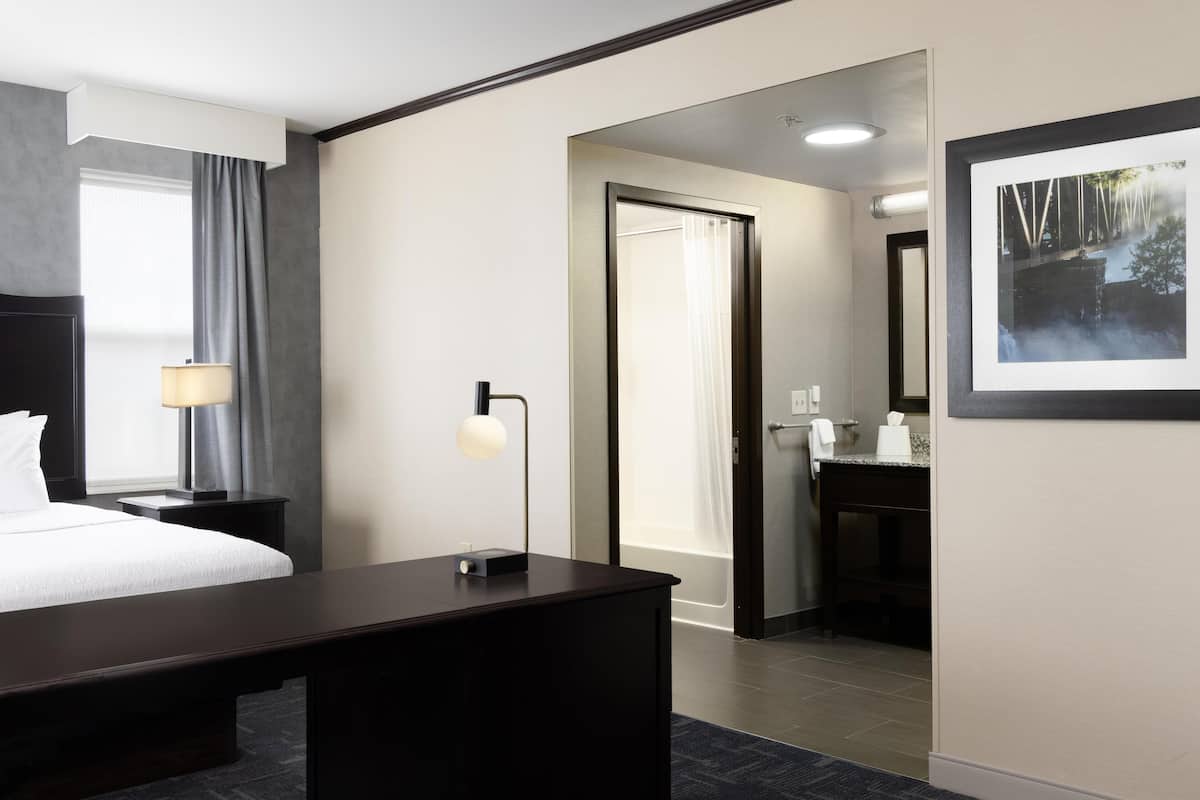 A hotel room with a bed, bedside table, lamp, and a desk. There is a bathroom visible with a mirror, sink, and towel rack.
