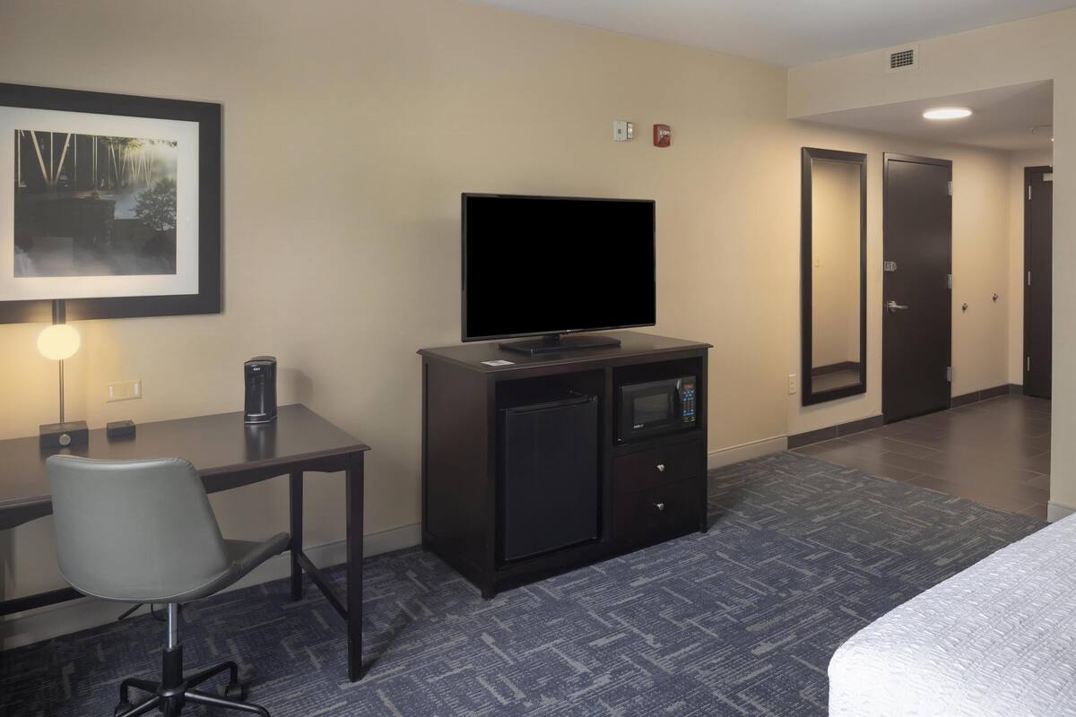 A hotel room with a flat-screen TV, a small refrigerator, a microwave, a desk with a chair, and a bed.