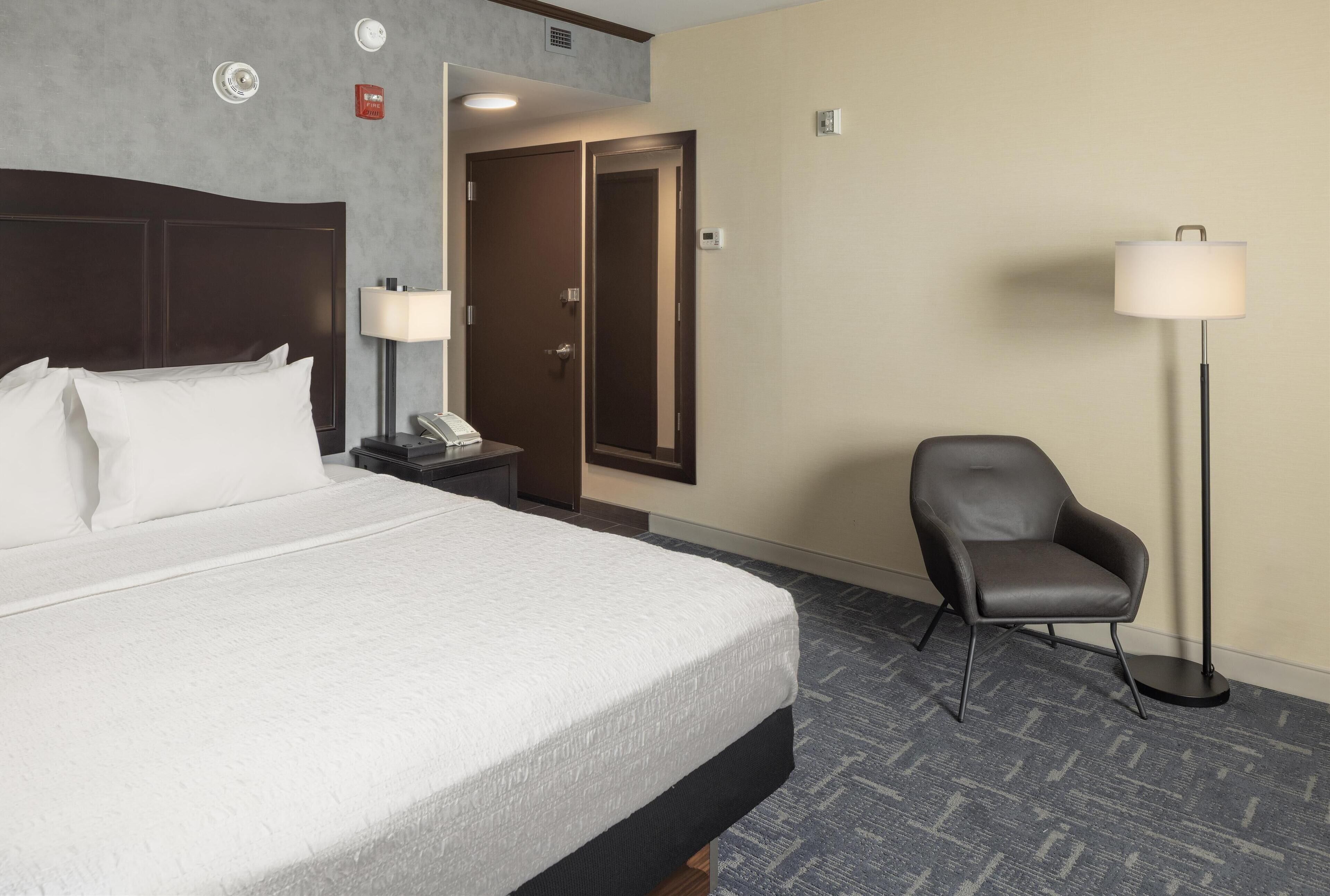 Een hotelkamer met een groot bed, een stoel, een bureau met een lamp en een deur naar een andere kamer.
