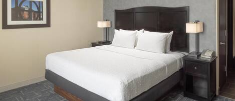 Quarto de hotel com uma cama grande, duas mesas de cabeceira com abajures, um quadro na parede e piso coberto por carpete.