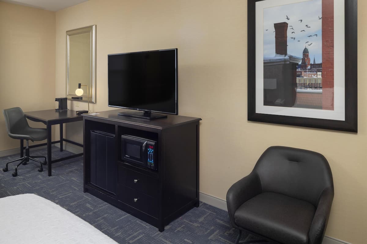 A hotel room with a bed, a desk with a chair, a TV, a microwave, and a framed picture on the wall.