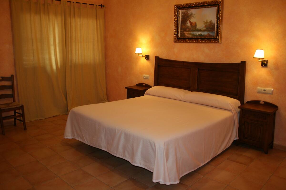 A neatly made bed with white linens, two bedside tables with lamps, a chair, and a painting on the wall.