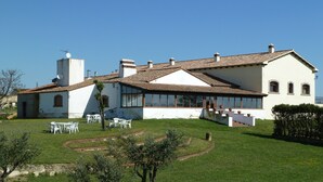 Un edificio blanco con techo de tejas, un pequeño patio con sillas blancas y un césped con árboles.