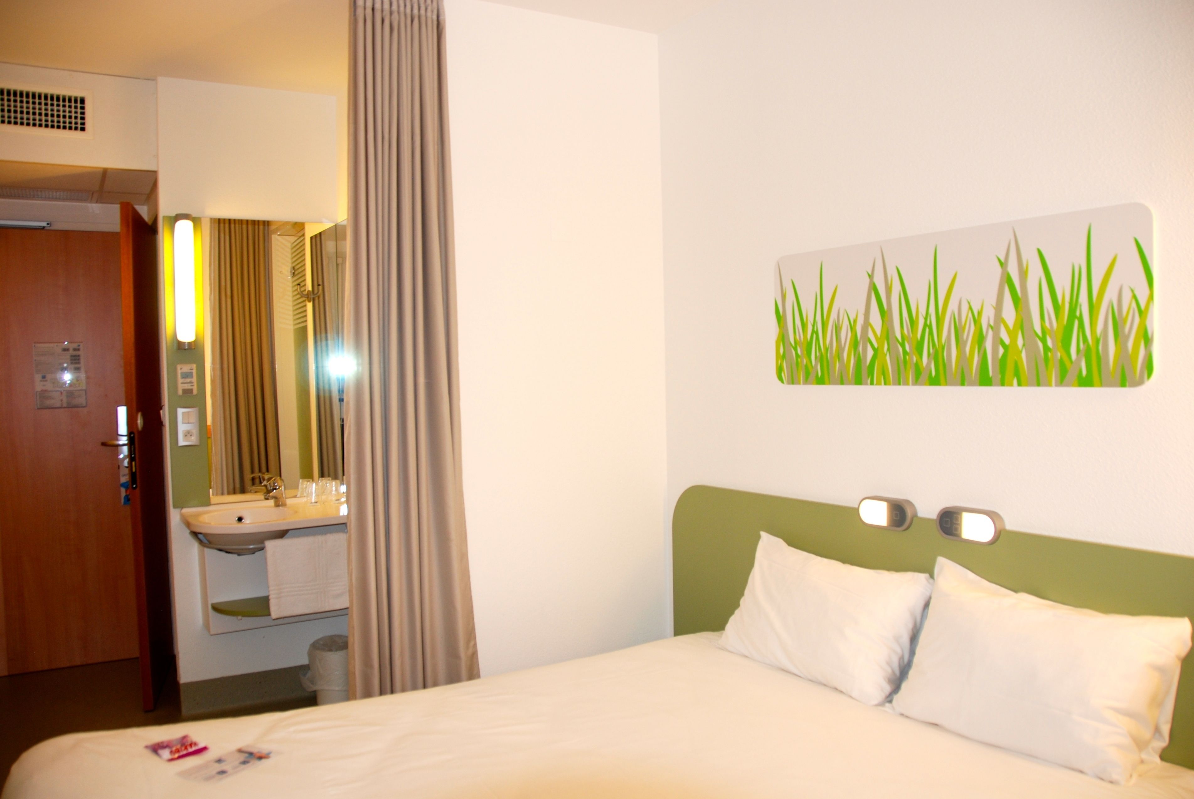 Une chambre d’hôtel avec un lit, une tête de lit verte, une lampe fixée au mur et une salle de bain visible à travers une porte ouverte.