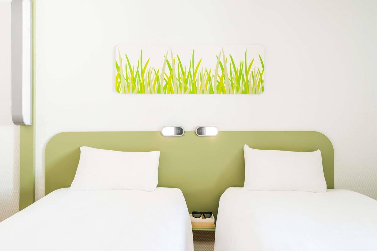 A hotel room with a green headboard, two white beds, a wall-mounted light, and a wall art of grass.