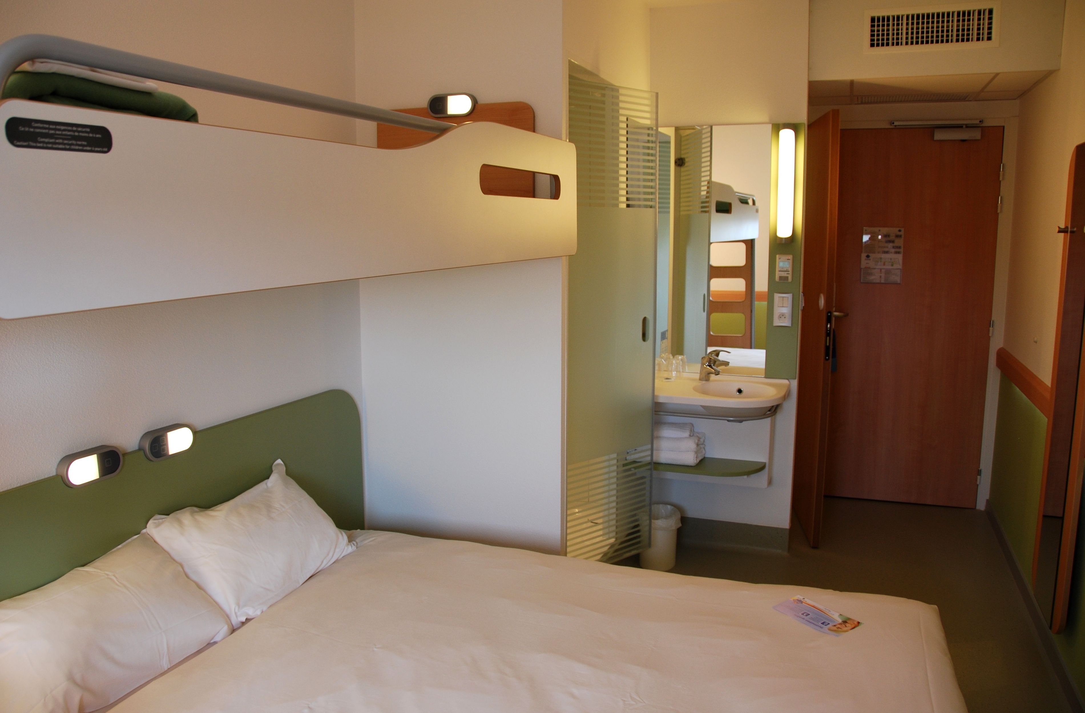 A hotel room with a bunk bed, a single bed, a bathroom with a sink and mirror, and a door leading to another room.