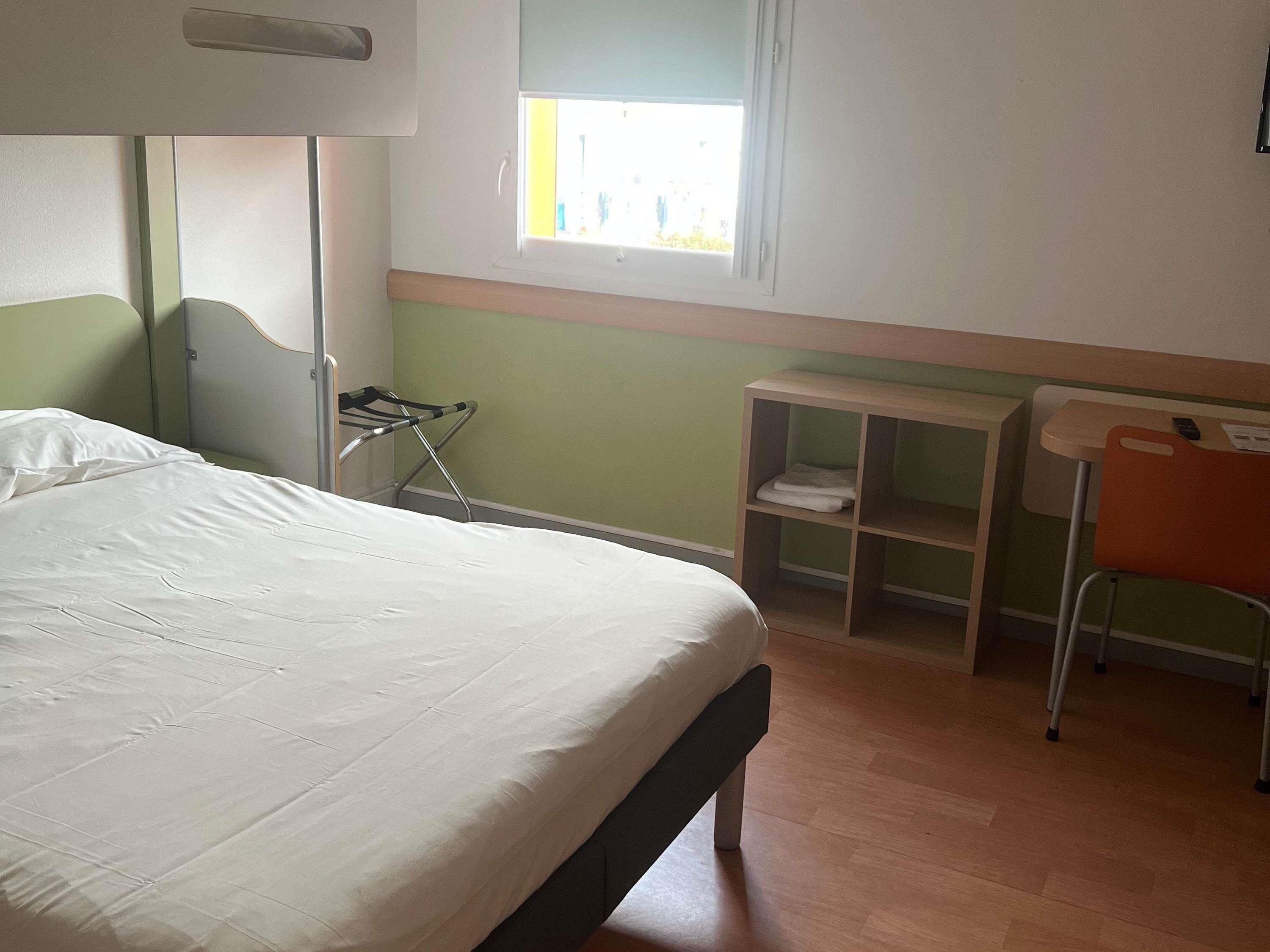 A single bed with a headboard, a small table with a chair, a shelf with folded towels, and a window with daylight.