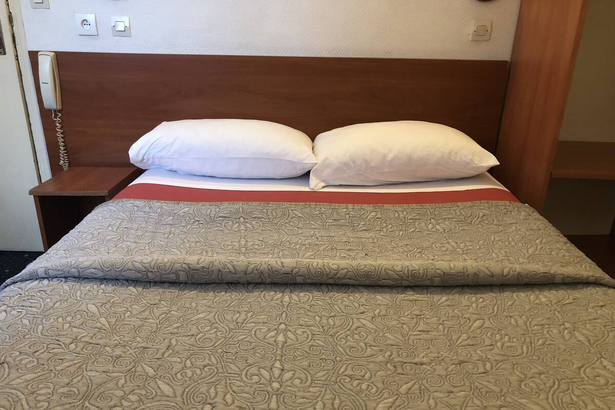 Un lit bien fait, recouvert d’une couvre-lit à motifs et agrémenté de deux oreillers, une table de chevet en bois avec un téléphone, et un mur équipé de prises électriques.