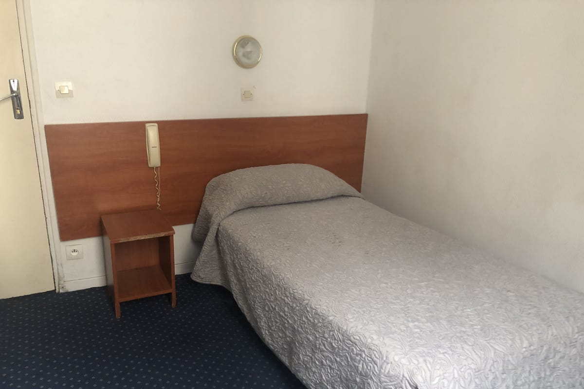 Un lit simple avec une tête de lit en bois, une table de chevet et un téléphone fixé au mur.