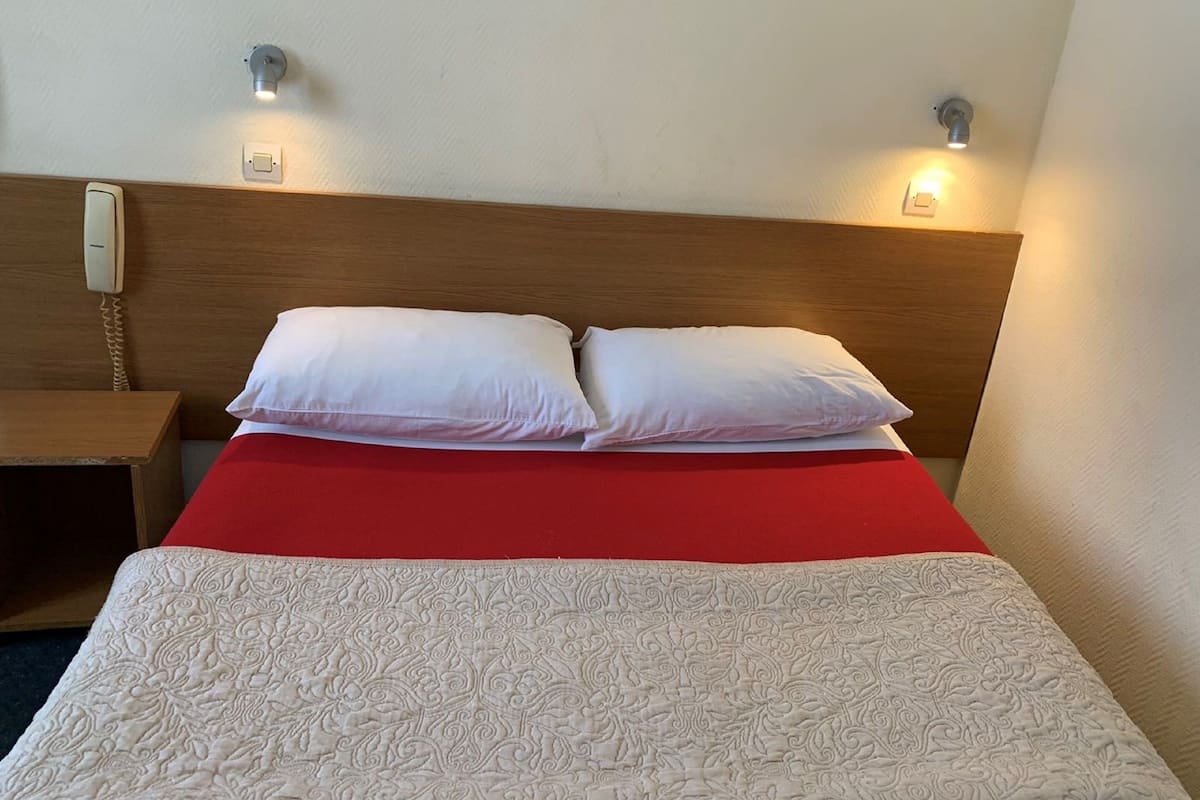 Une chambre d’hôtel avec une tête de lit en bois, deux lampes de chevet, un téléphone, une couvre-lit rouge et une housse de couette blanche matelassée.