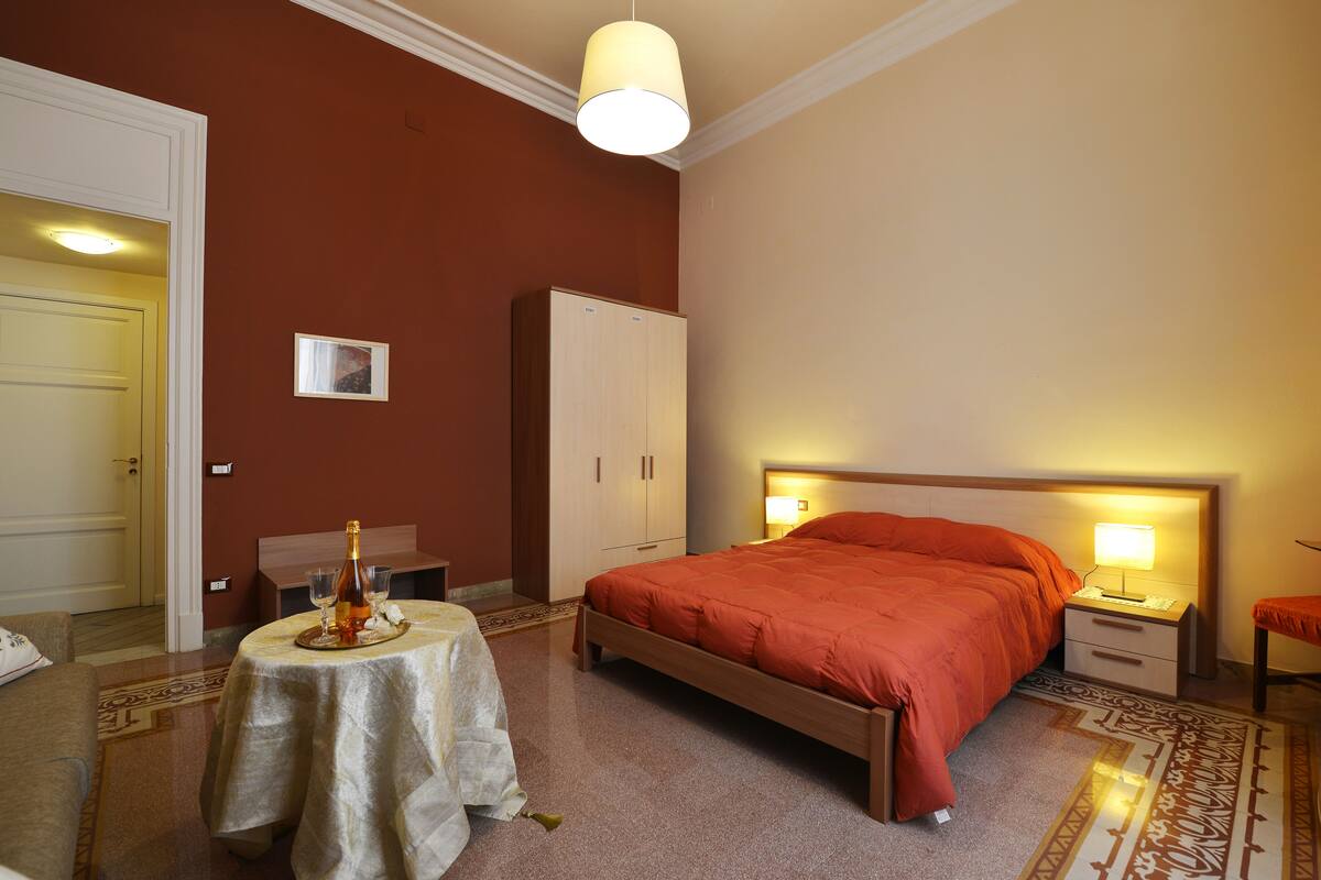 A hotel room with a bed, bedside table, lamp, wardrobe, and a small table with a bottle and glasses.
