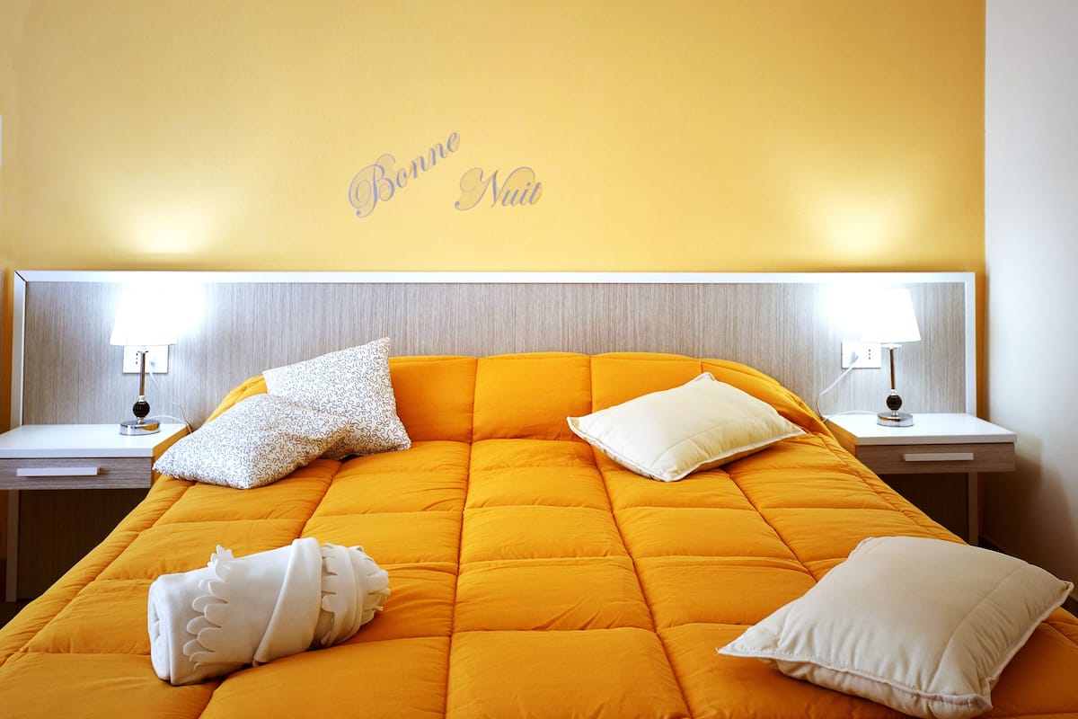 A hotel room with a large orange bed, two bedside lamps, and a wall with the text "Bonne Nuit".