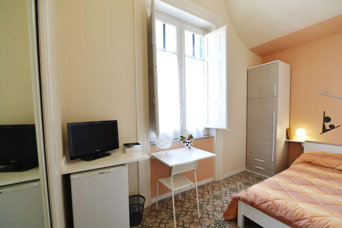A bedroom with a bed, a desk with a computer, a wardrobe, and a window with curtains.