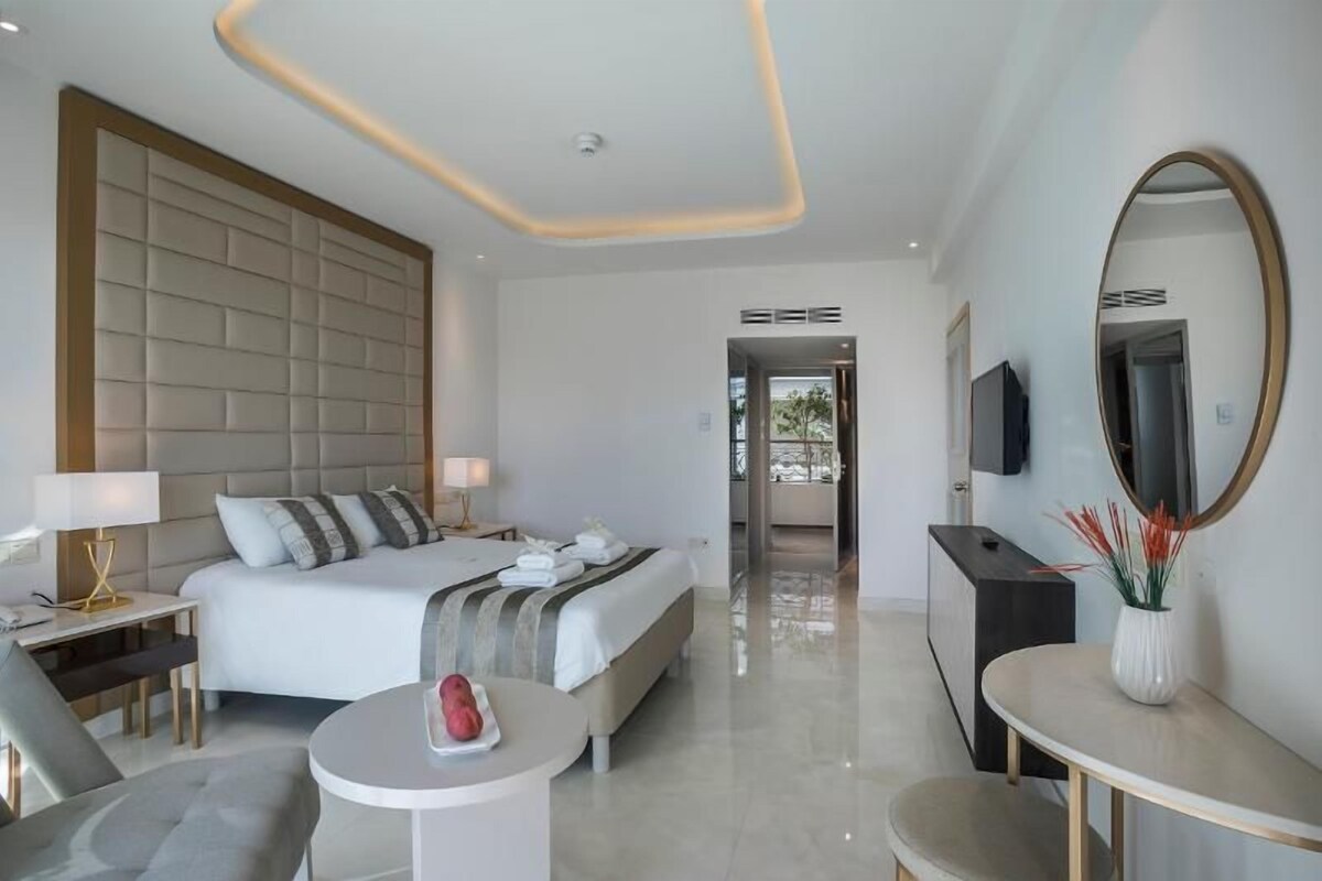 A modern hotel room with a large bed, bedside tables, a round coffee table, a TV, and a mirror.